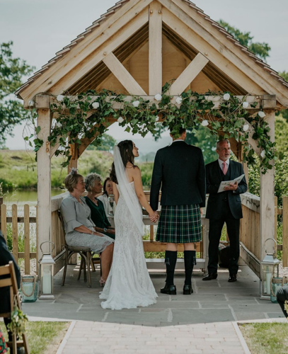 A bride and groom under a wooden canopy next to a river. They are facing away from the camera.